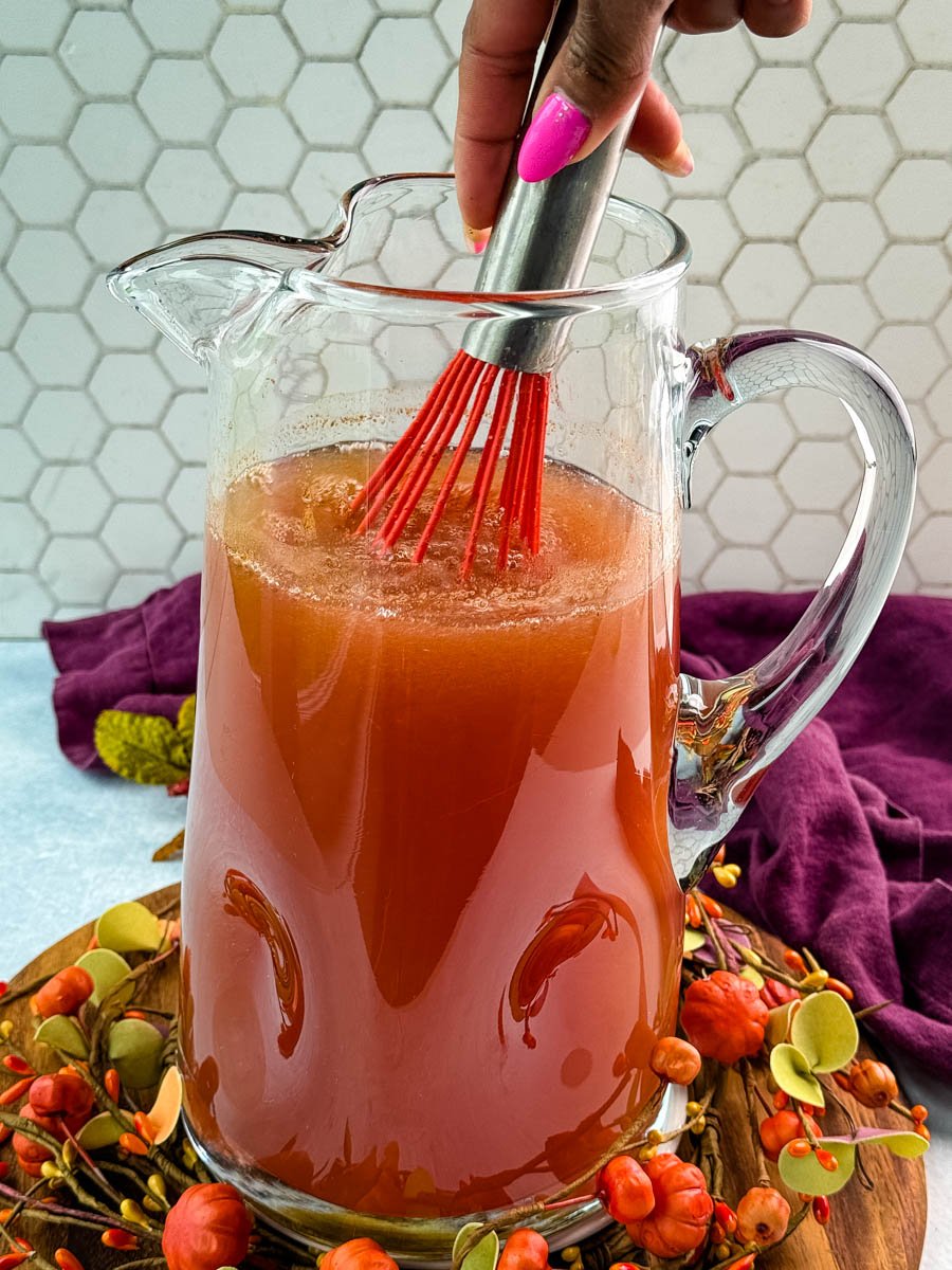 Hand using a red whisk to stir a pitcher of Thanksgiving punch, blending the spiced mixture