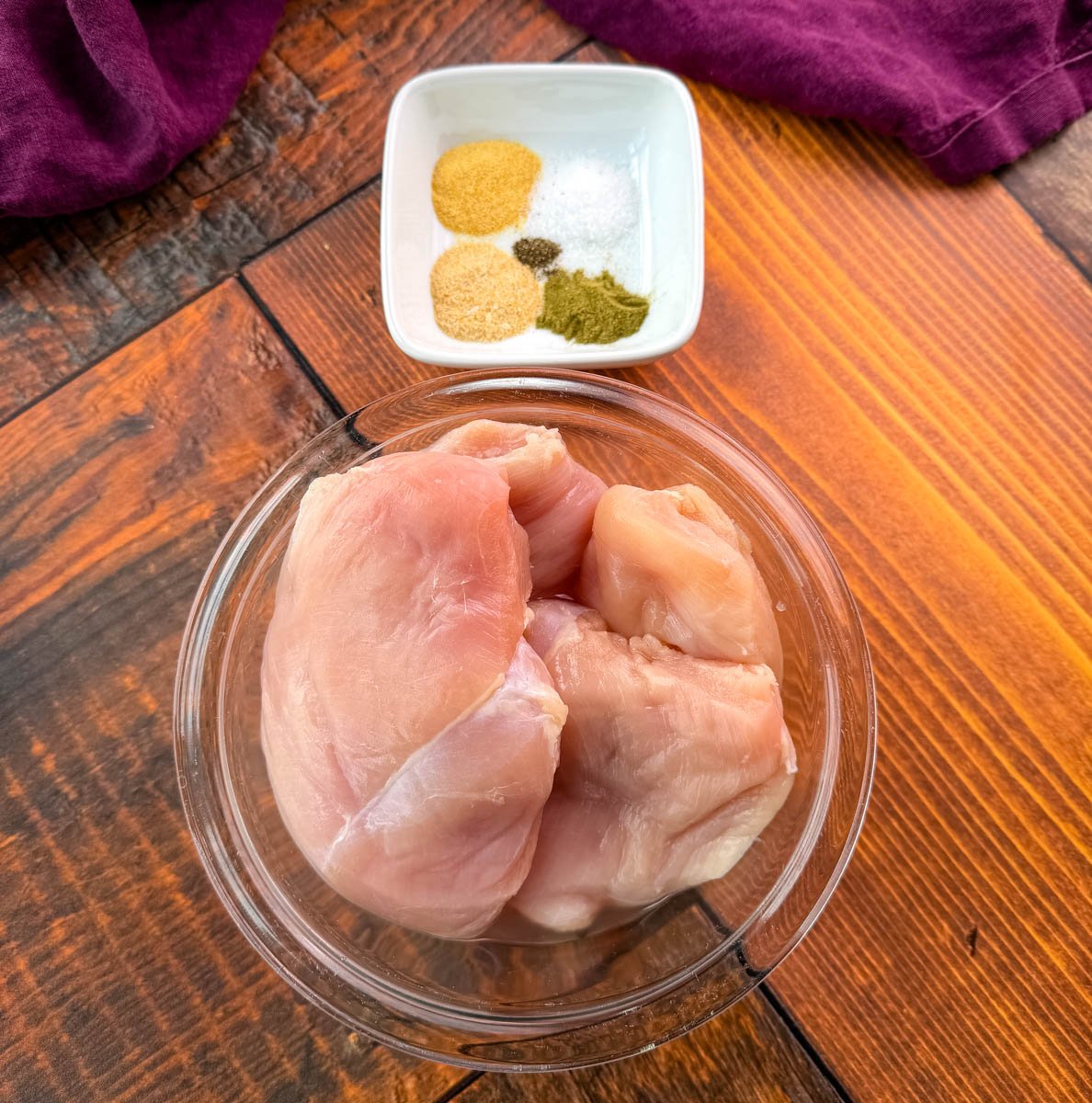 A clear bowl filled with raw chicken breasts on a wooden surface, with a small square dish of seasonings including garlic powder, onion powder, salt, pepper, and herbs.