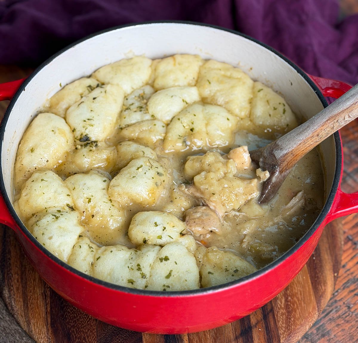 A red Dutch oven filled with chicken and dumplings, topped with fluffy biscuit dumplings sprinkled with herbs, with a wooden spoon resting inside.