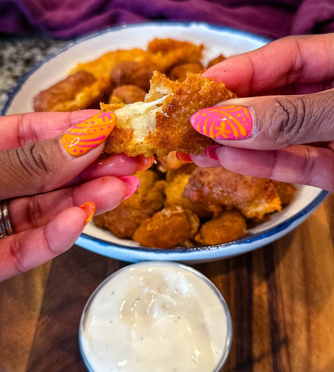 Close-up of hands pulling apart a crispy fried cheese curd to reveal gooey melted cheese inside, with a bowl of ranch dipping sauce below.