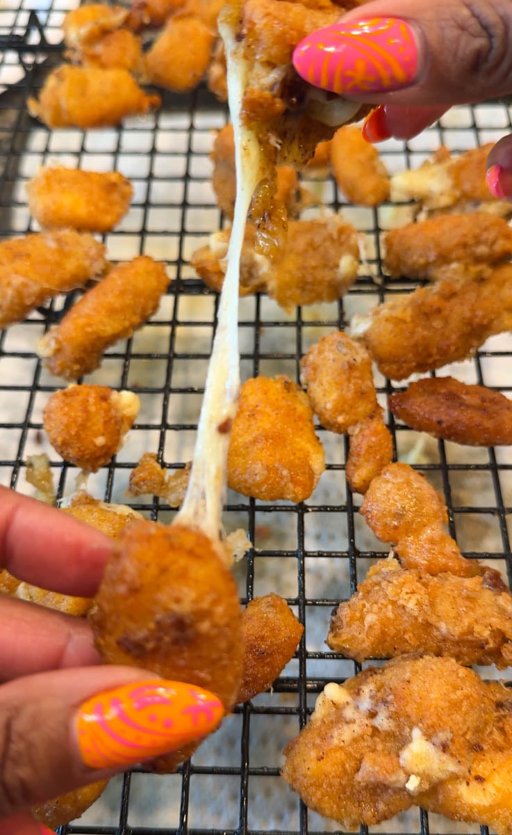 Close-up of golden fried cheese curds on a cooling rack with gooey melted cheese stretching between two pieces being pulled apart by hand.