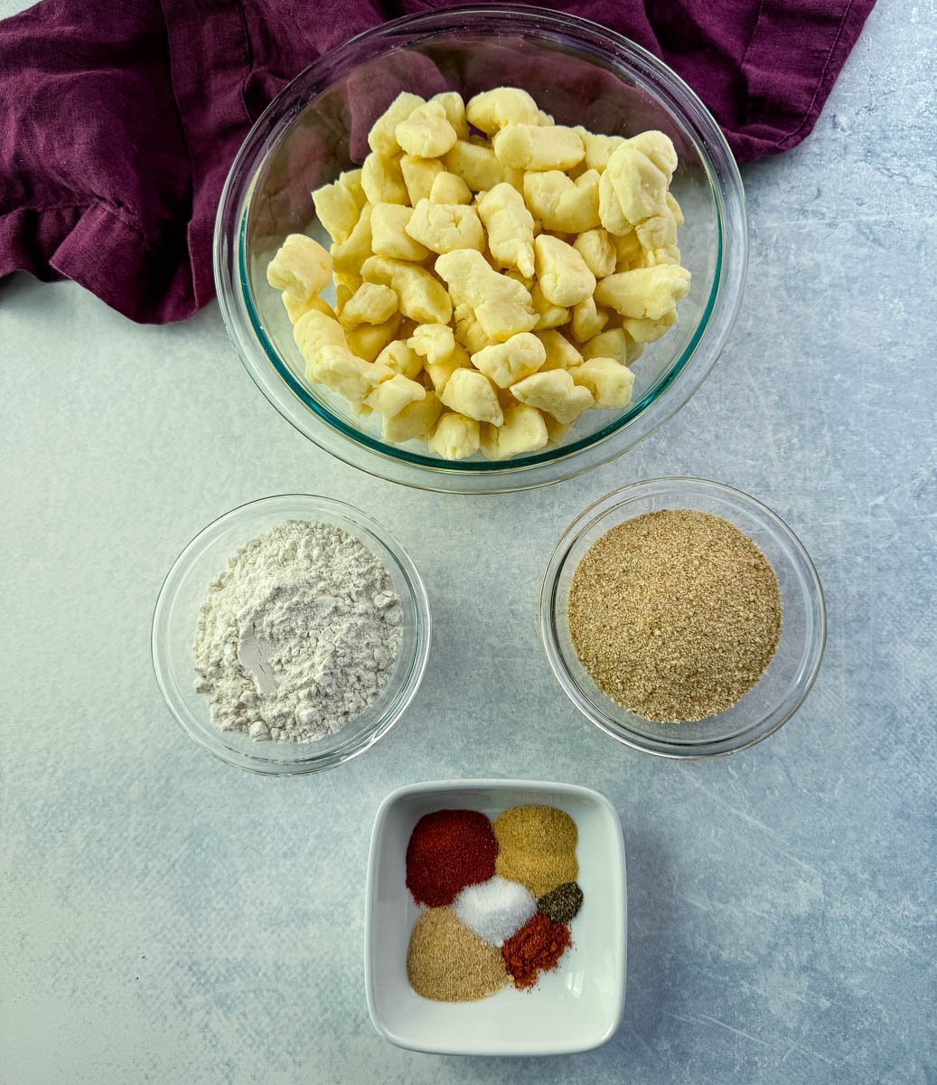 Overhead view of cheese curd ingredients in bowls: fresh cheese curds, flour, breadcrumbs, and a mix of colorful spices on a light background.