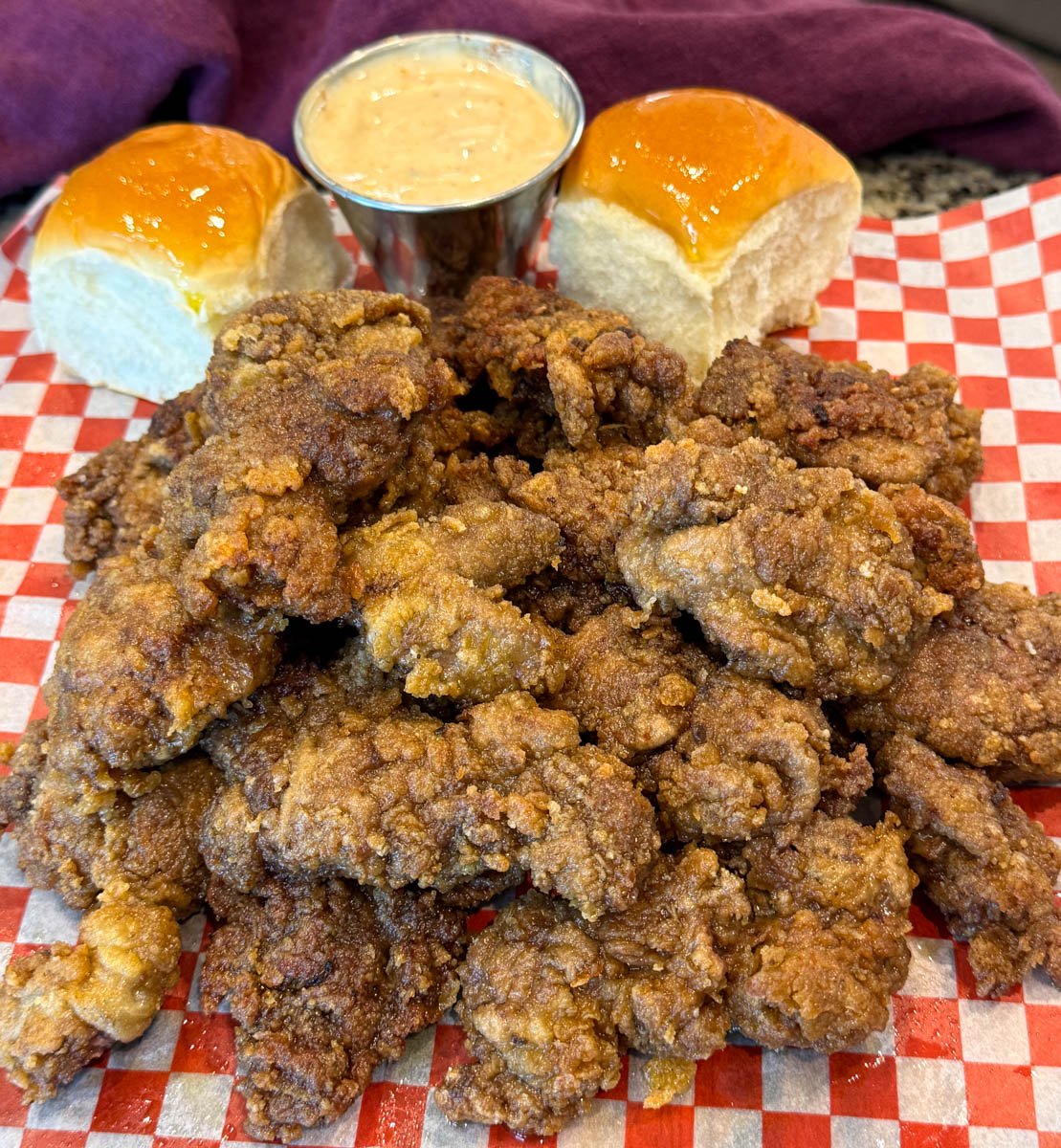 Crispy fried chicken livers stacked on checkered paper, paired with dinner rolls and a dipping sauce.