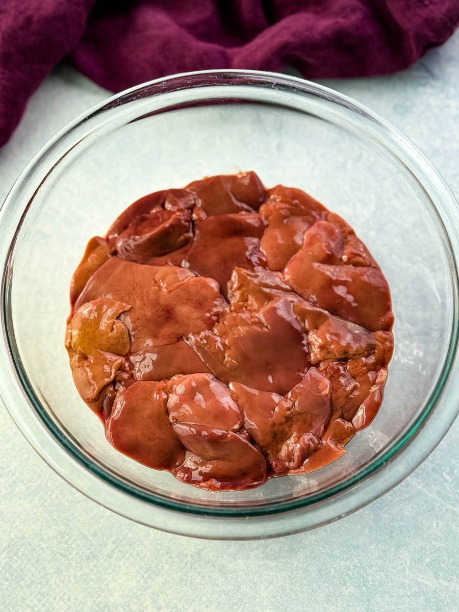 A clear bowl filled with raw chicken livers, set on a light surface with a deep purple cloth in the background.