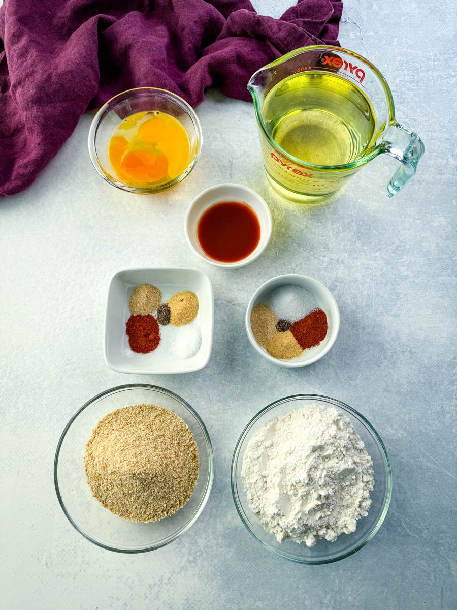 Eggs, hot sauce, flour, breadcrumbs, seasonings, and oil measured out in bowls and a measuring cup, all arranged neatly on a light countertop.