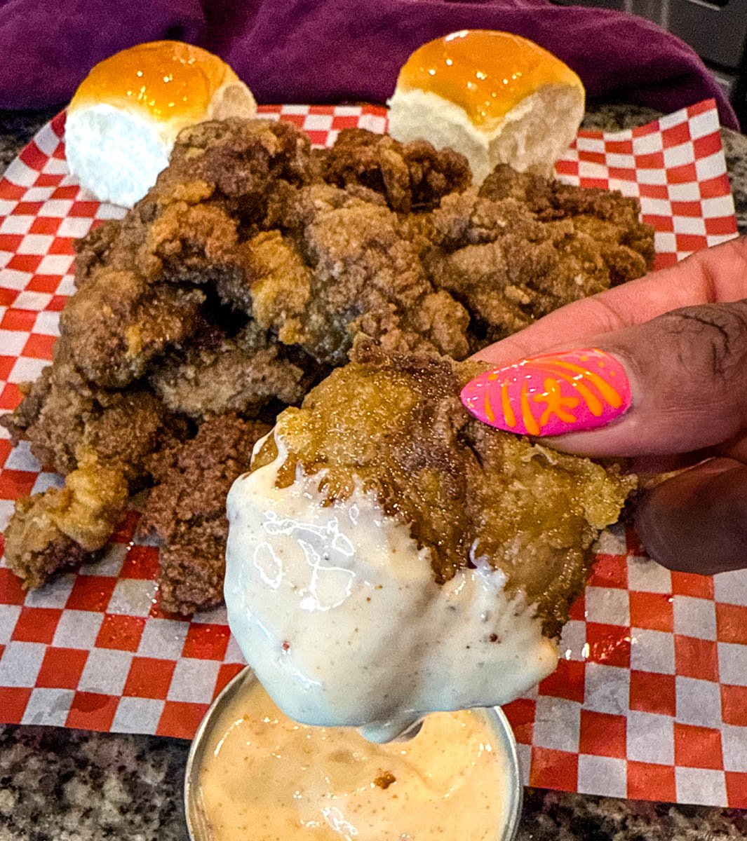 A hand holding a crispy fried chicken liver over a small metal cup of dipping sauce, with a pile of fried livers in the background.