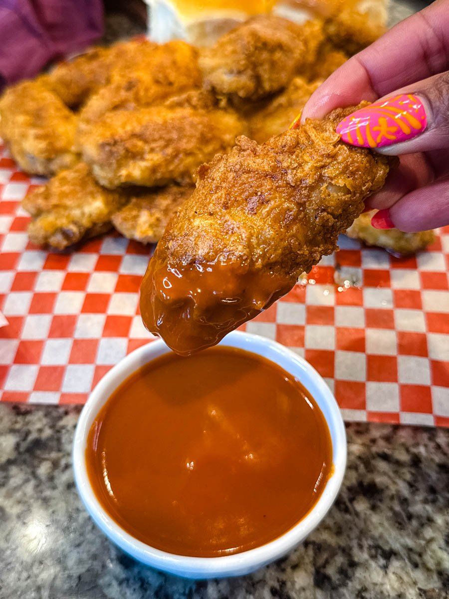 Hand holding a crispy fried chicken wing dipped into a bowl of bright orange buffalo sauce, with more fried wings in the background.