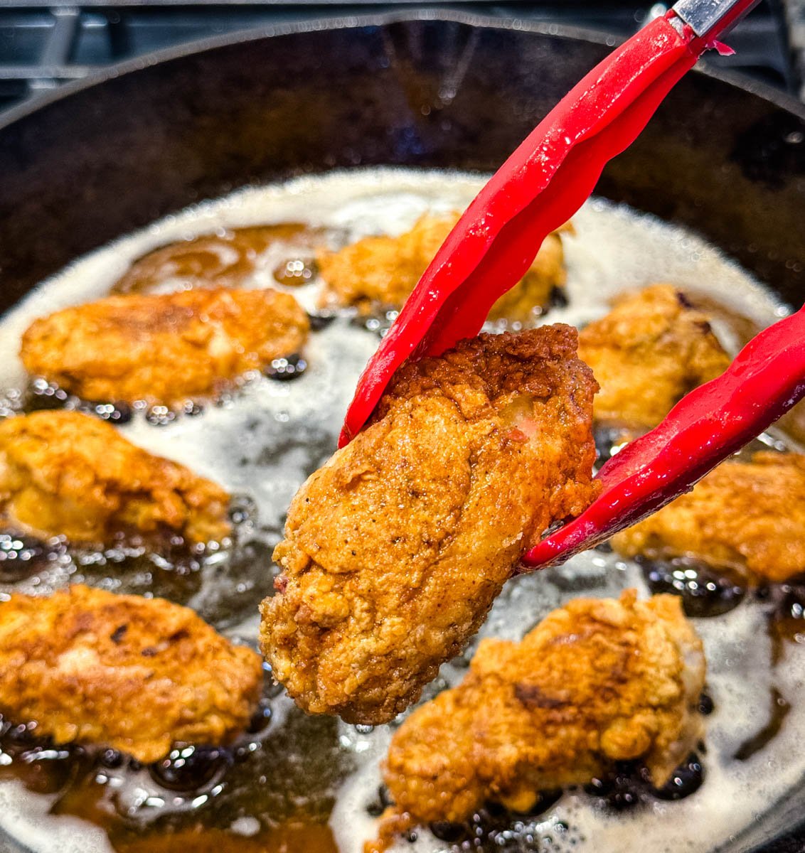 Crispy fried chicken wing held with red tongs over a skillet of bubbling hot oil with several pieces frying.