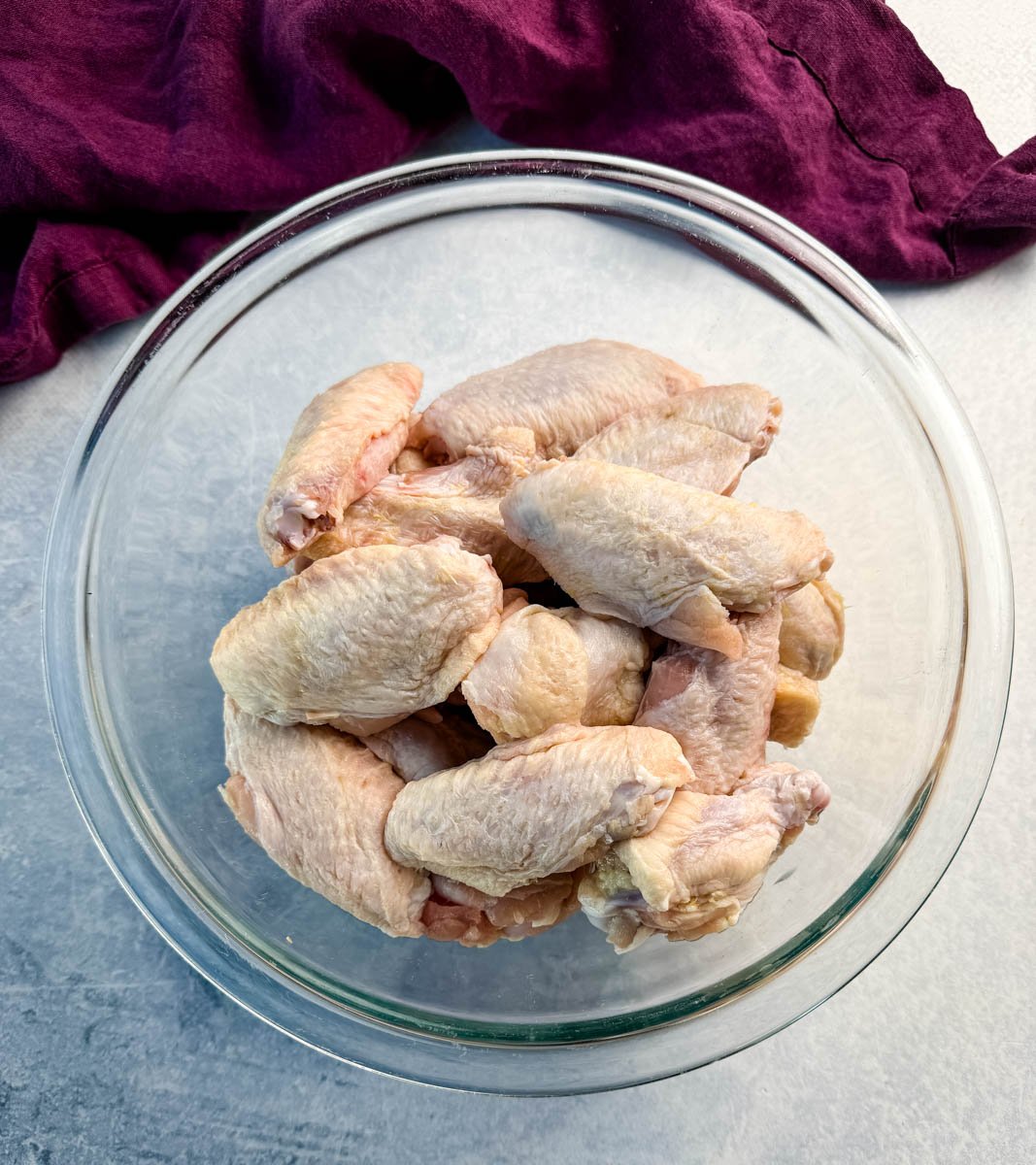 A clear glass bowl filled with raw chicken wings, placed on a counter with a purple cloth in the background.