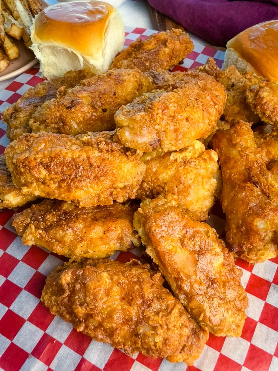Crispy fried chicken wings stacked high on red-and-white checkered paper with fluffy dinner rolls beside them.
