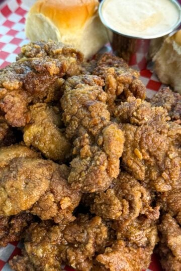 A generous serving of crispy fried chicken livers piled high on a tray lined with checkered paper, with rolls and sauce on the side.