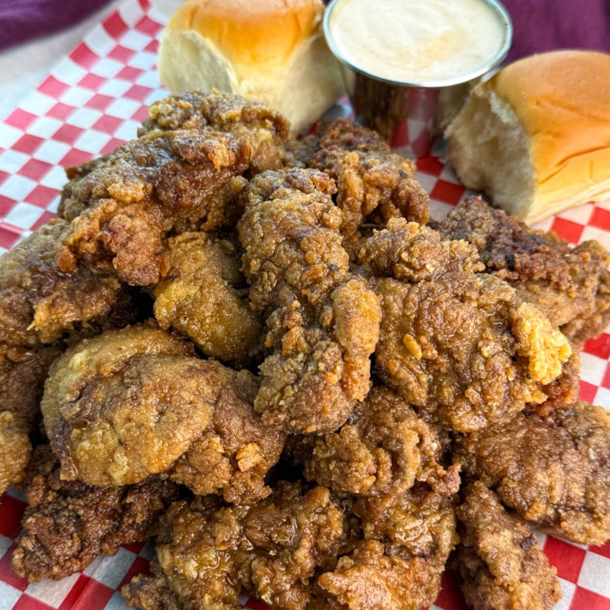 A generous serving of crispy fried chicken livers piled high on a tray lined with checkered paper, with rolls and sauce on the side.