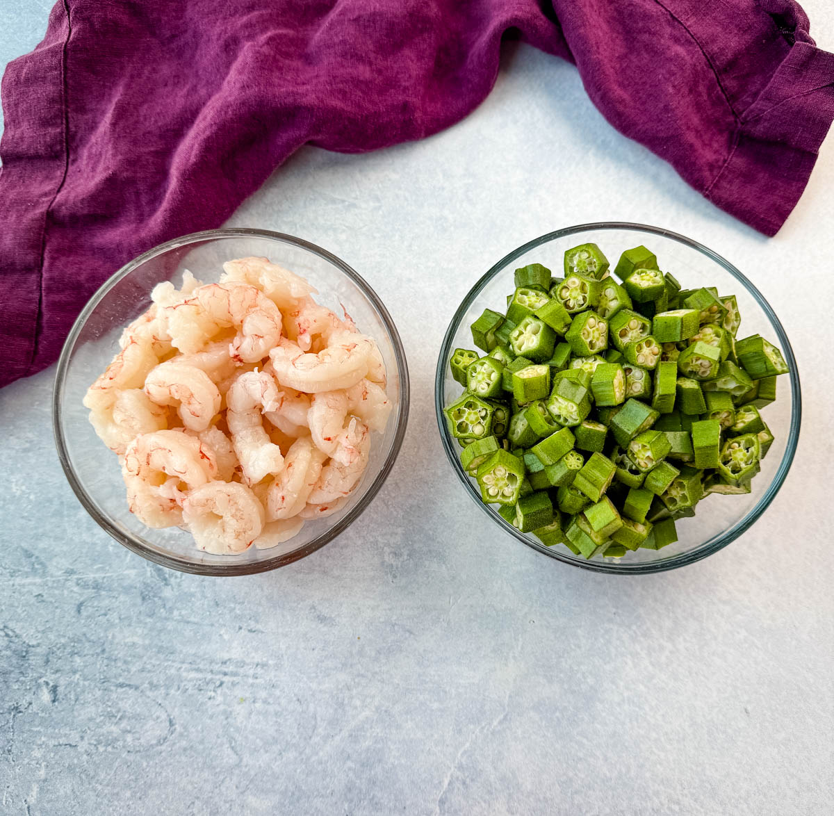 Two glass bowls side by side—one filled with raw peeled shrimp, the other with freshly chopped okra. A purple kitchen towel is in the background.