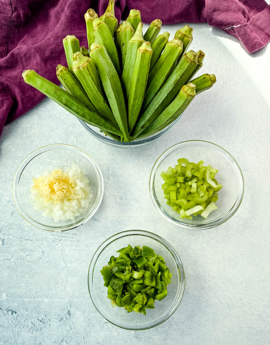 Fresh whole okra next to bowls of diced onion with garlic, chopped celery, and green bell pepper.