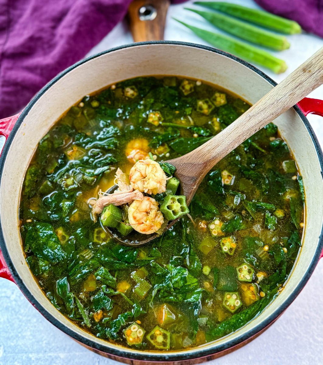Wooden spoon lifting shrimp, okra, and spinach from a simmering pot of okra soup in seasoned broth.