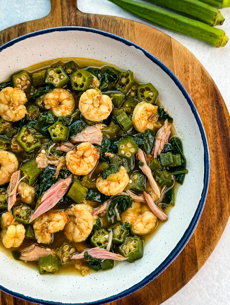 Bowl of okra soup with shrimp, smoked turkey pieces, and collard greens in broth, served on a wooden board with fresh okra nearby.