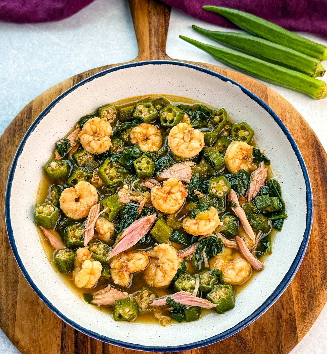 Overhead view of a bowl of okra soup filled with shrimp, okra, and shredded smoked turkey, placed on a wooden board with whole okra to the side.