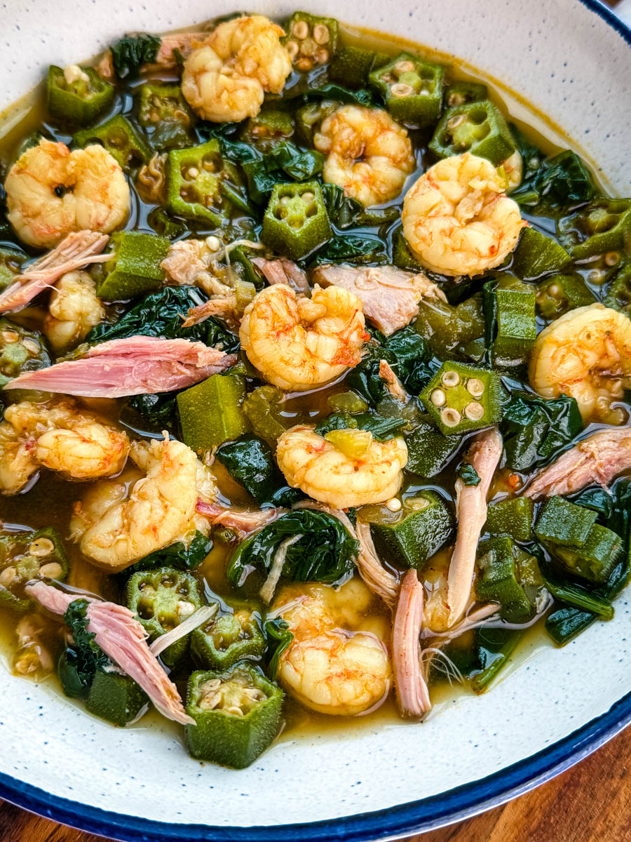 Close-up of okra soup showing shrimp, tender okra, shredded smoked turkey, and wilted greens in a rich broth.