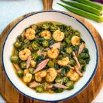 Bowl of okra soup filled with shrimp, chunks of smoked turkey, spinach, and okra in a rich broth, served on a wooden board with fresh okra in the background.