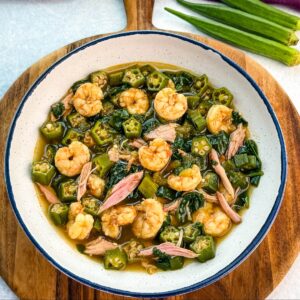 Bowl of okra soup filled with shrimp, chunks of smoked turkey, spinach, and okra in a rich broth, served on a wooden board with fresh okra in the background.