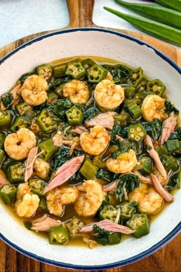 Bowl of okra soup filled with shrimp, chunks of smoked turkey, spinach, and okra in a rich broth, served on a wooden board with fresh okra in the background.