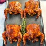 Four golden-brown baked Cornish hens arranged on a black roasting pan, garnished in the center with a sprig of fresh rosemary.