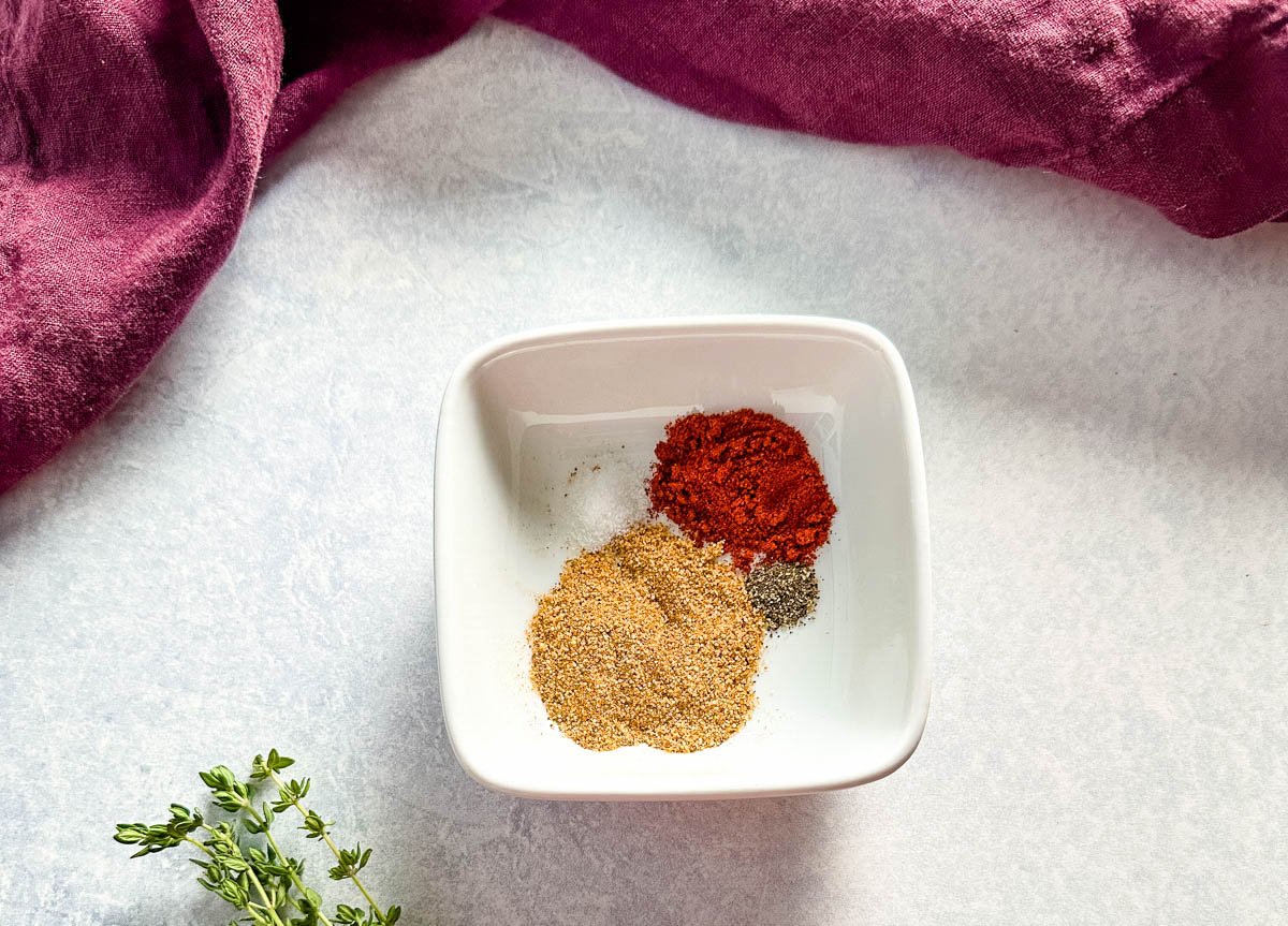 A small white dish filled with a spice blend, including paprika, black pepper, and garlic powder, with a sprig of fresh thyme nearby on the counter.