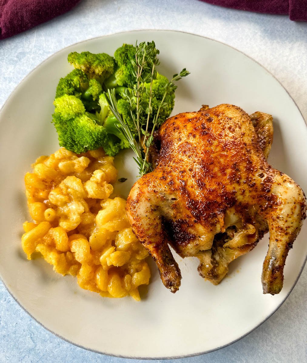 A plated serving with one baked Cornish hen alongside bright green broccoli florets and creamy baked macaroni and cheese, garnished with fresh thyme.