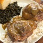 Plate of turkey chops smothered in brown gravy over rice, served with collard greens and a dinner roll.