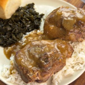 Plate of turkey chops smothered in brown gravy over rice, served with collard greens and a dinner roll.
