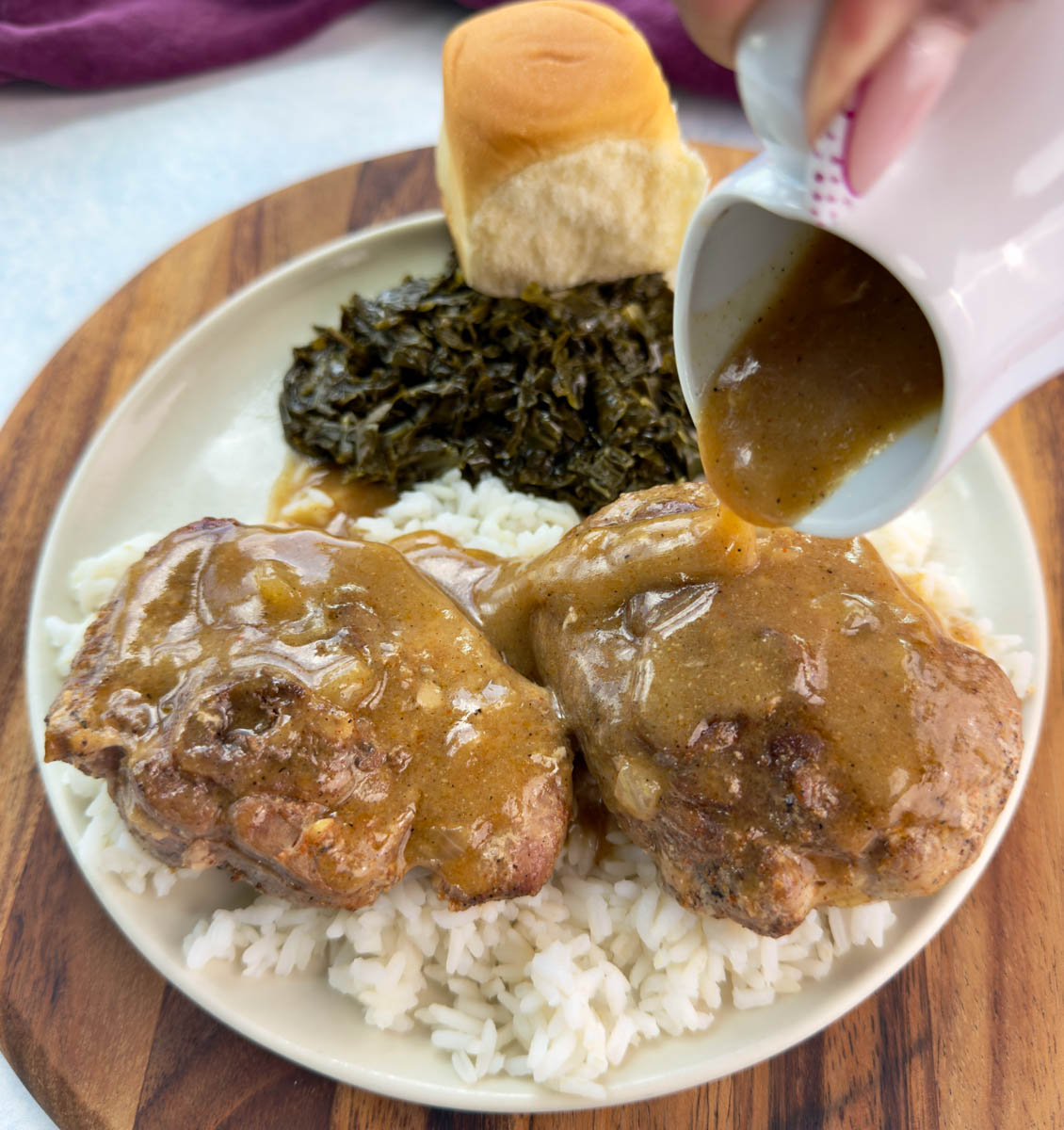 Close-up of gravy being poured from a white pitcher over turkey chops served with rice, collard greens, and a roll.