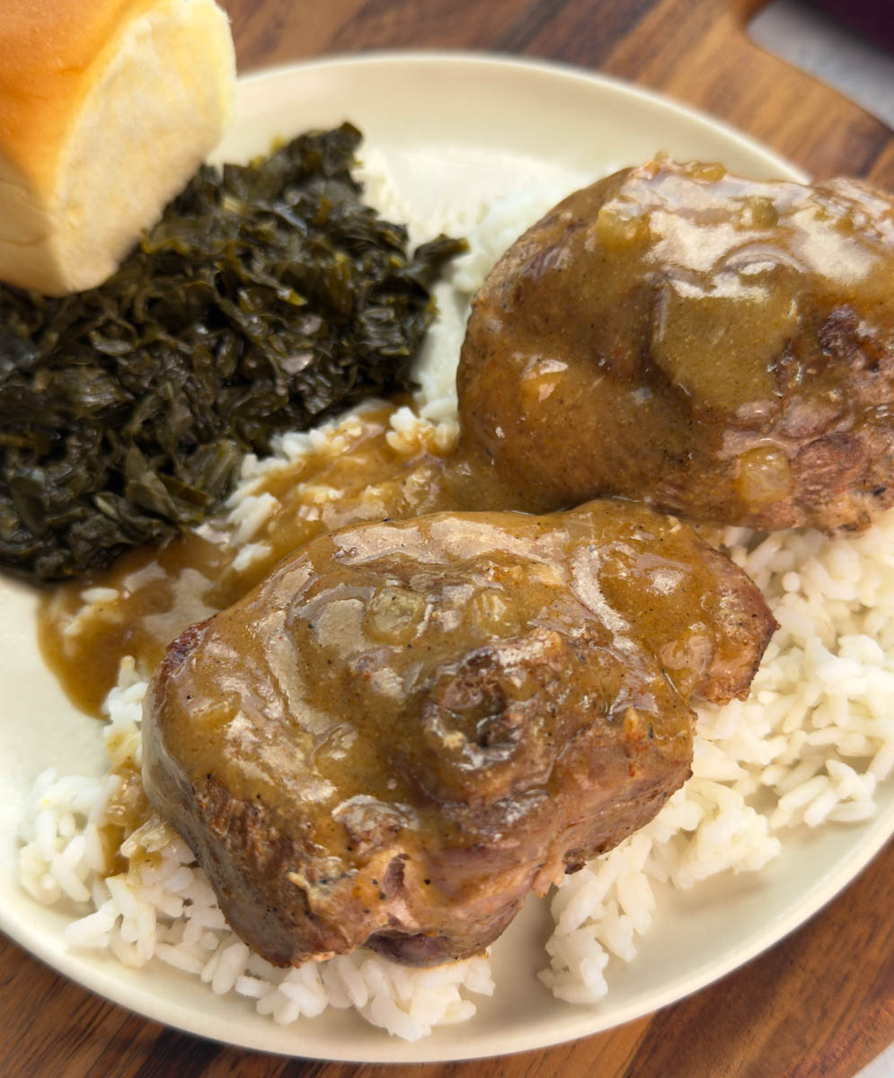 Plate of turkey chops smothered in brown gravy over rice, served with collard greens and a dinner roll.