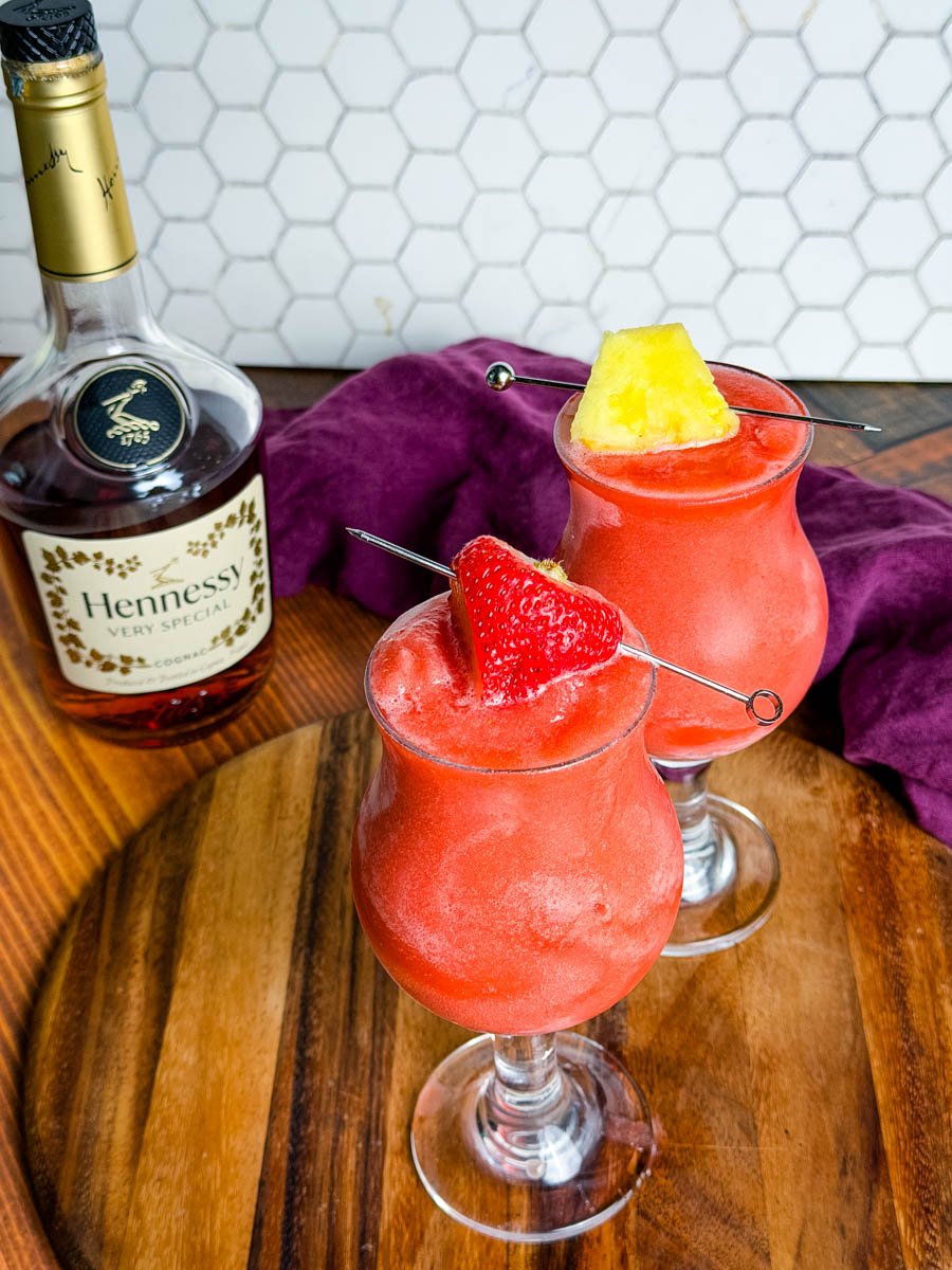 Two bright red Hennessy slushies in curved hurricane glasses topped with a strawberry and pineapple slice, sitting on a wooden board with a bottle of Hennessy and a purple napkin in the background.