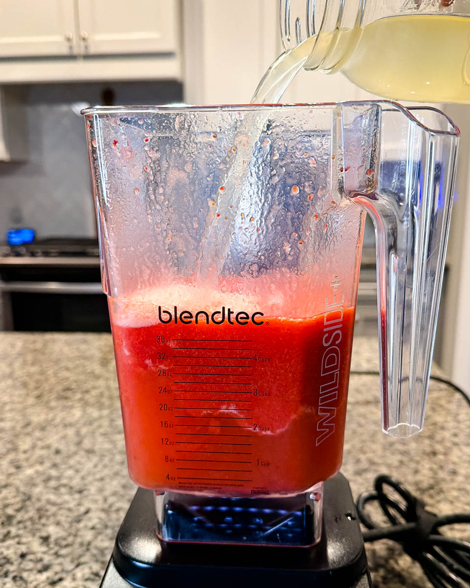 Blendtec blender with frozen strawberry mixture inside as pineapple juice is being poured in to prep for the Hennessy slushie.