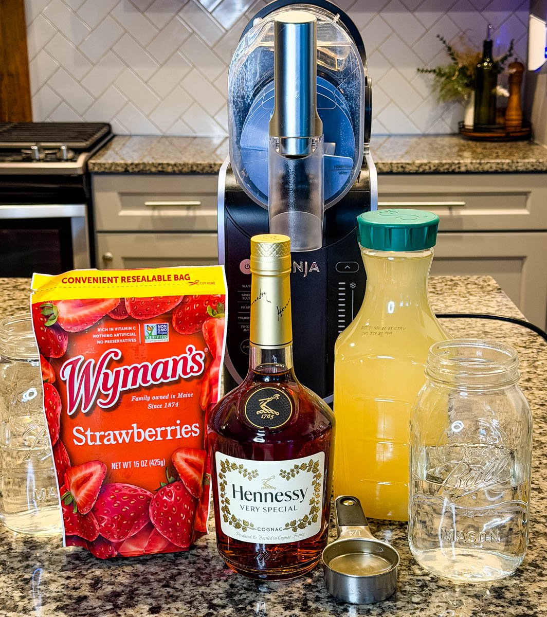 Ingredients lined up on a kitchen counter including frozen strawberries, pineapple juice, water, a bottle of Hennessy, and a Ninja Slushi machine in the background.