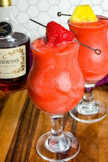 Two bright red Hennessy slushies in curved hurricane glasses topped with a strawberry and pineapple slice, sitting on a wooden board with a bottle of Hennessy and a purple napkin in the background.