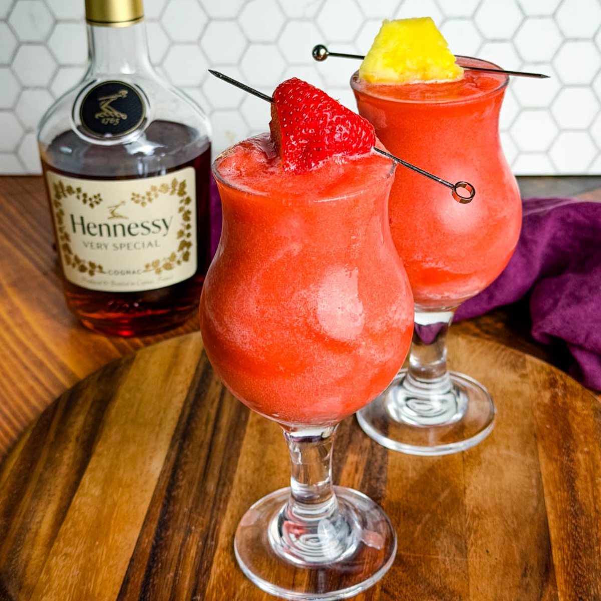 Two bright red Hennessy slushies in curved hurricane glasses topped with a strawberry and pineapple slice, sitting on a wooden board with a bottle of Hennessy and a purple napkin in the background.