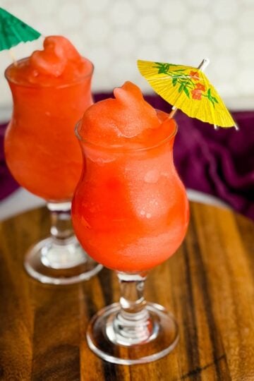Two glasses of frozen Ninja Slushi Rum Punch topped with mini paper umbrellas on a wooden board.