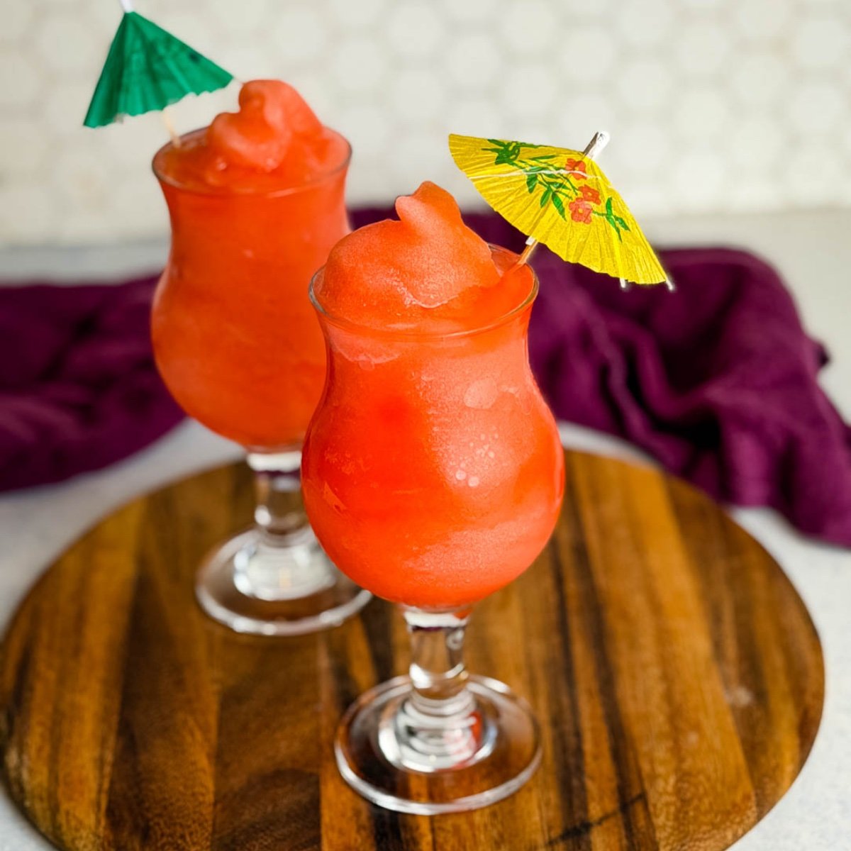 Two glasses of frozen Ninja Slushi Rum Punch topped with mini paper umbrellas on a wooden board.