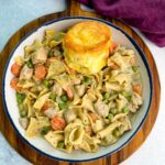 Close-up of a creamy one-pot chicken pot pie casserole with egg noodles, peas, and carrots served in a cast-iron skillet topped with golden biscuits.