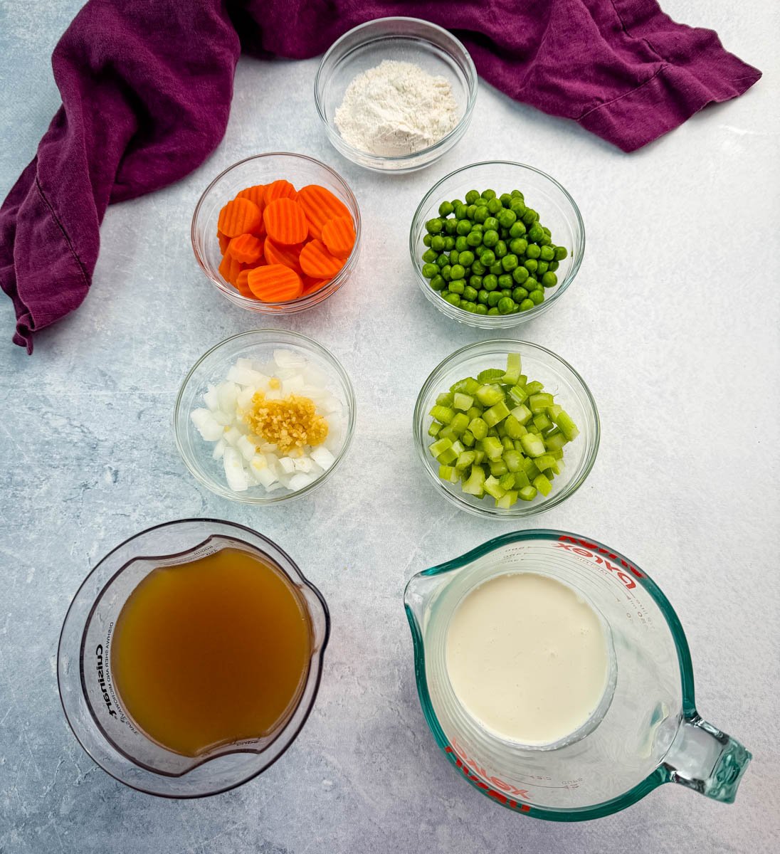 Bowls of prepped ingredients for chicken pot pie casserole — carrots, peas, celery, onion with garlic, flour, chicken broth, and cream — arranged on a gray surface with a deep purple kitchen towel in the background.