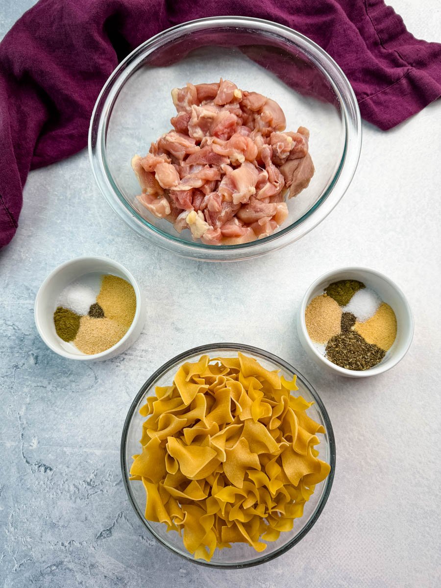Ingredients for chicken pot pie casserole laid out, including a bowl of raw chicken pieces, uncooked egg noodles, and two small bowls of seasonings, set on a light countertop with a purple cloth napkin.