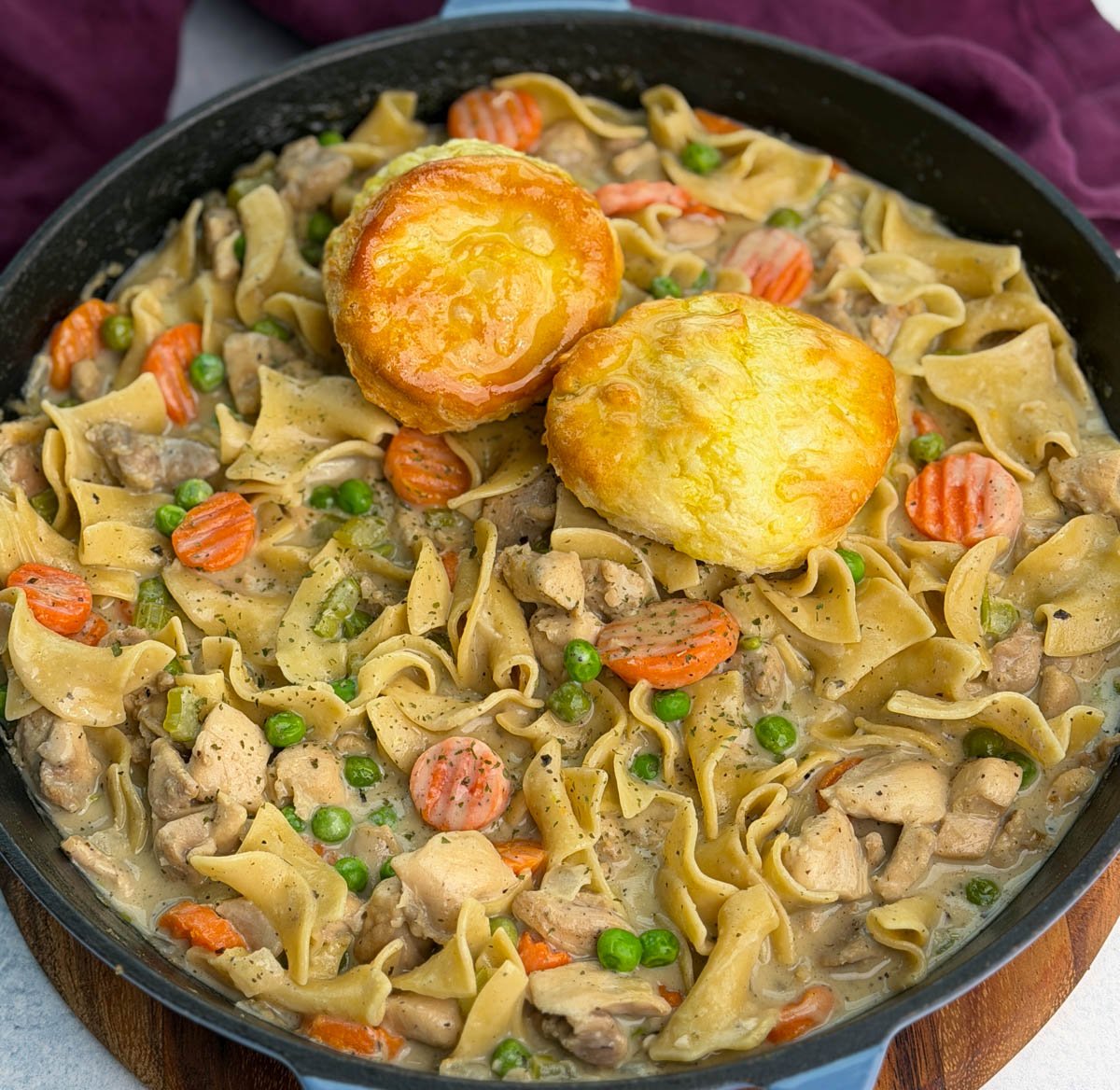 Skillet full of creamy chicken pot pie casserole with tender noodles, carrots, peas, and chicken, topped with golden biscuits for serving.