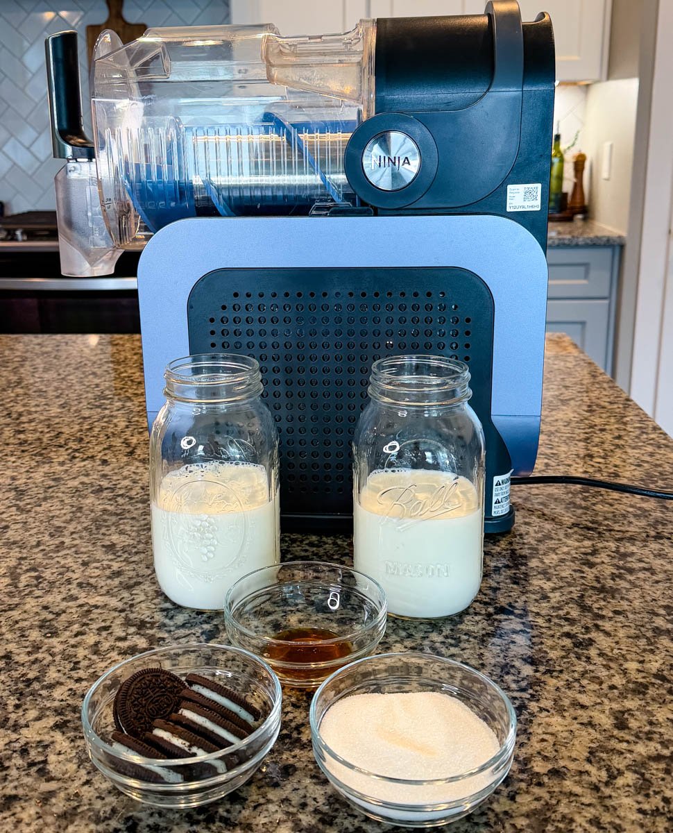 Ninja Slushi machine on a kitchen counter with ingredients for a McFlurry, including two jars of milk, vanilla extract, sugar, and Oreo cookies in small glass bowls.