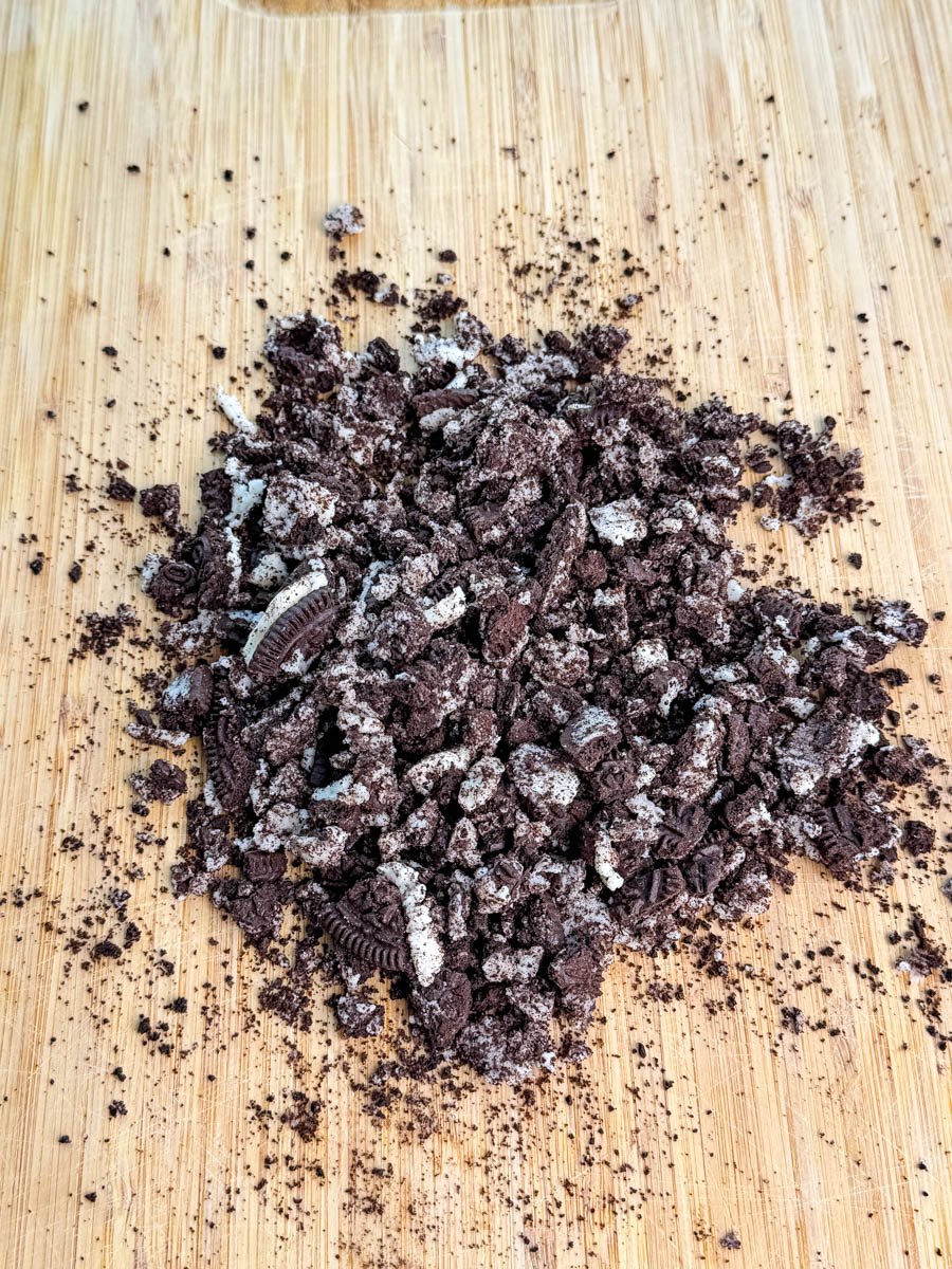 Crushed Oreo cookies on a wooden cutting board, showing cookie crumbs and cream filling pieces.