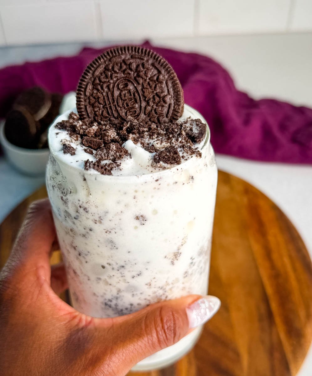 A hand holding a glass filled with a creamy Oreo Ninja Slushi McFlurry topped with crushed cookies and a whole Oreo cookie.
