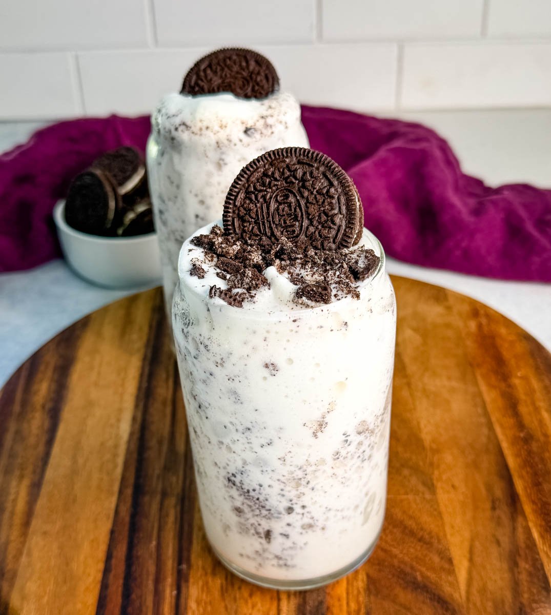 A tall glass of Oreo McFlurry-style Ninja Slushi topped with crushed cookies and a whole Oreo on top, sitting on a wooden board with a purple cloth in the background.