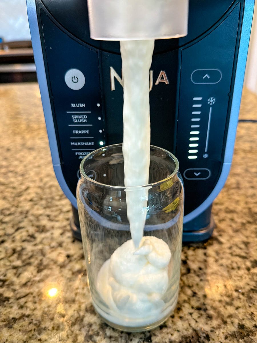 A creamy vanilla mixture dispensing from a Ninja Slushi machine into a clear glass on a granite countertop, showing the thick McFlurry-style texture forming as it pours.
