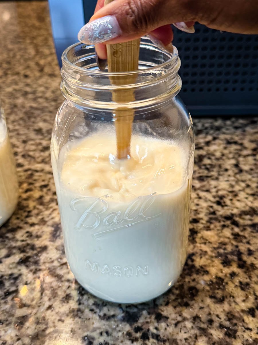A hand stirring a creamy milk mixture inside a mason jar on a granite countertop before freezing for the Ninja Slushi McFlurry.