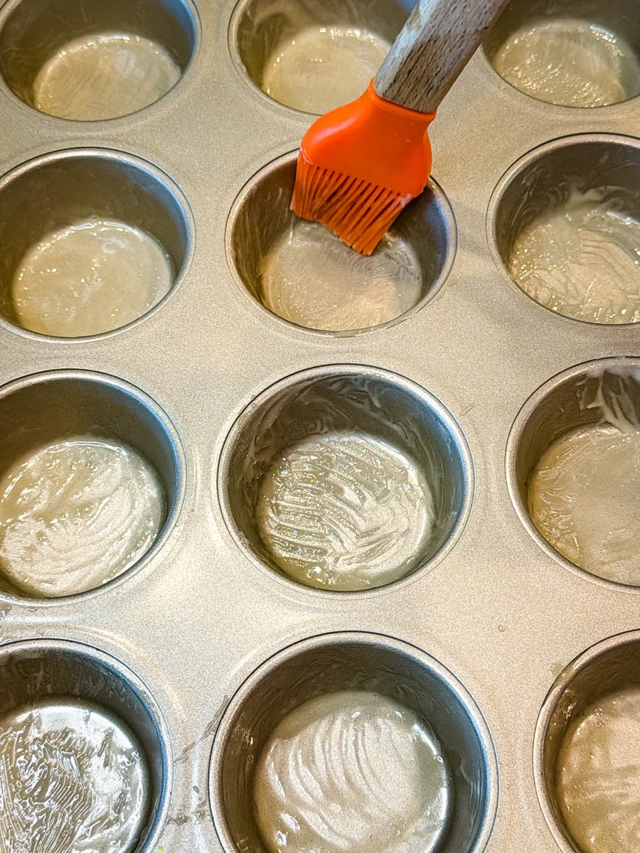 A muffin tin being greased with melted butter using an orange silicone brush, preparing the pan for baking cornbread muffins.